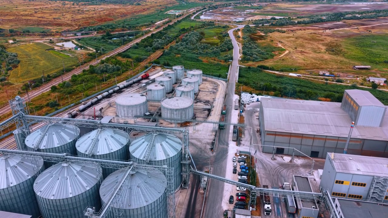 Getreideterminal mit Silos, Lagertanks und Bahngleisen. Schräge Drohnenansicht von Metallsilos, Flachdachtanks, Förderbändern, Fabrikgebäuden, Tankwaggons und Zufahrtsstraßen.