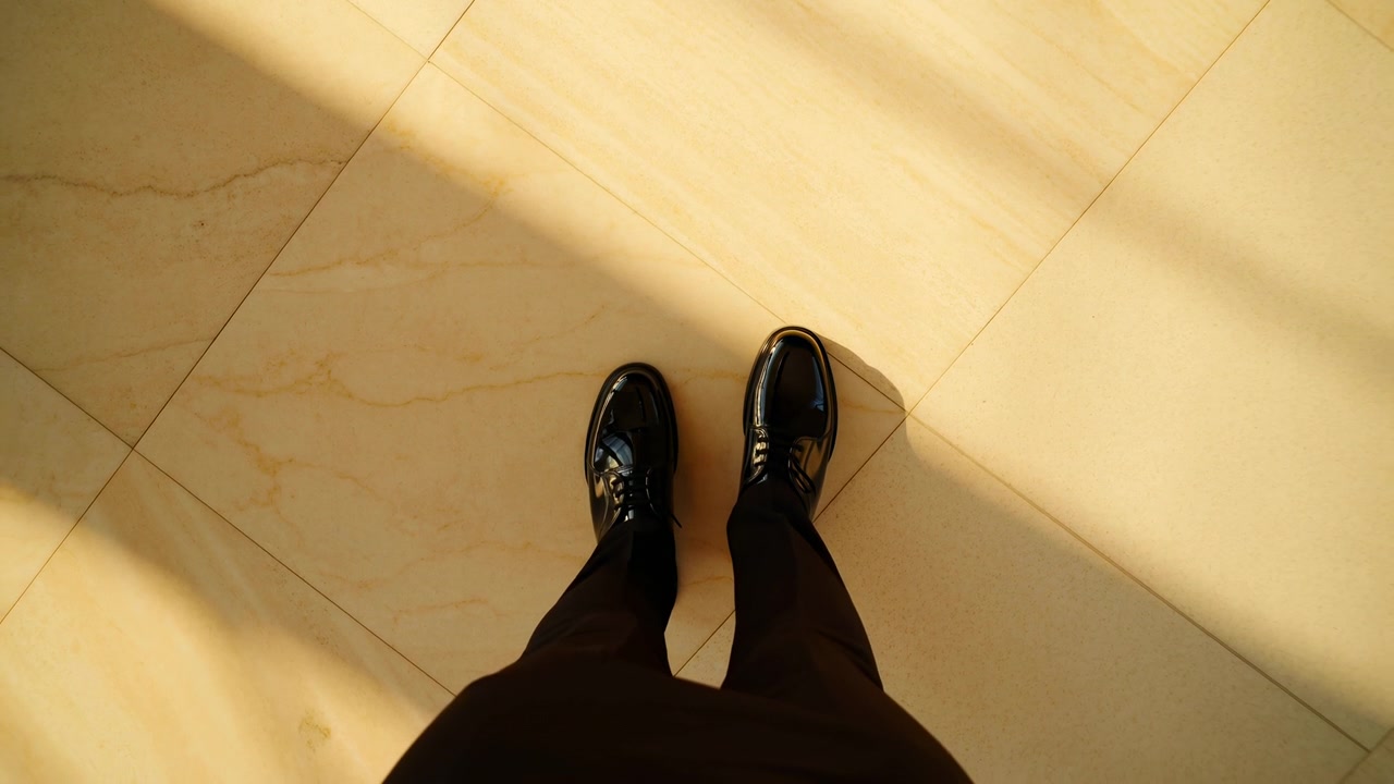 Un par de pies con zapatos negros permanece inmóvil sobre un suelo de baldosas en un ambiente interior neutro y tranquilo, iluminado por la luz del sol. La perspectiva cenital resalta la luz limpia y natural y el blanco