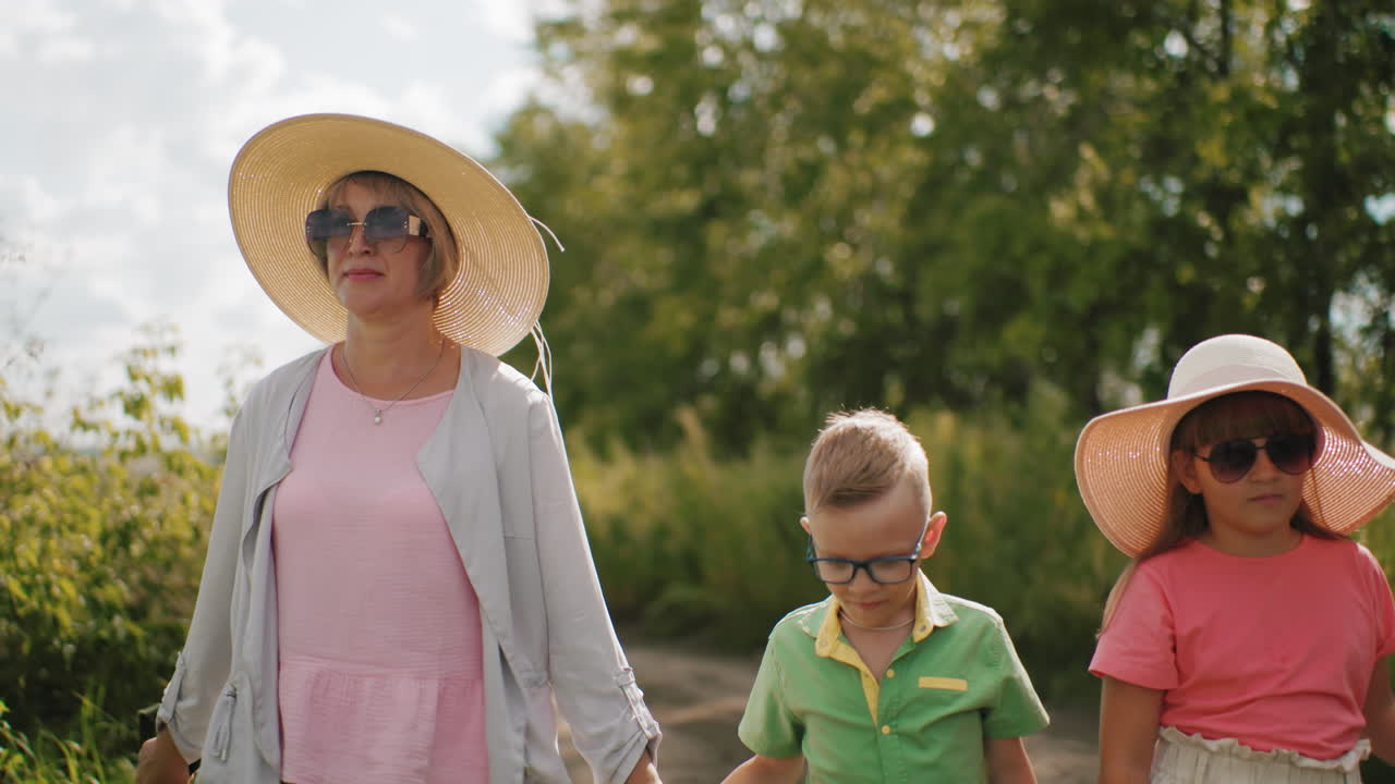 мать держит сына за руку во время прогулки по пышной зеленой тропе, дочь в темных солнцезащитных очках с серьезным выражением лица идет рядом с ними под ярким солнечным светом