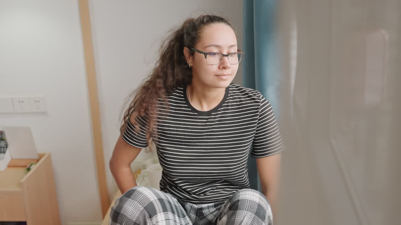 Donna seduta al finestrino di un hotel mentre prepara il tè, una scena mattutina in camera con lampada da comodino in legno, abiti a fantasia, tazza di ceramica, un rituale lento, umore contemplativo, viaggiatrice solitaria che controlla il telefono e si accomoda.