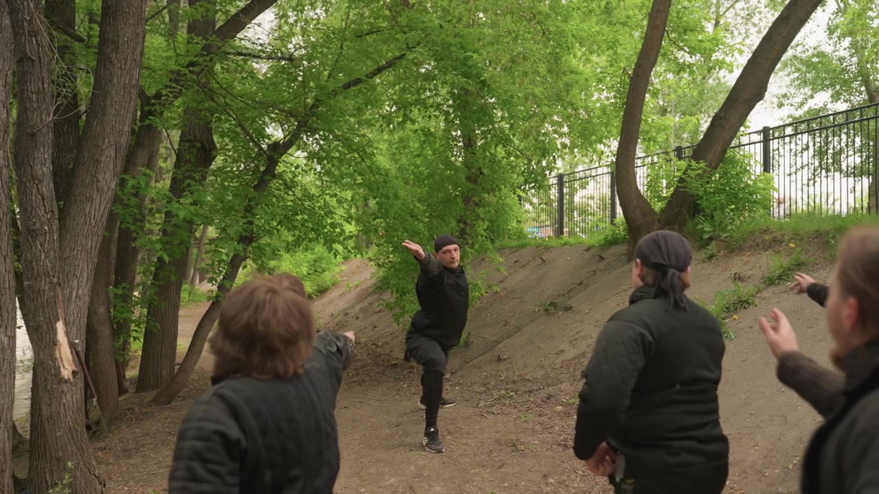 Group of men observe man demonstrating martial arts movements on dirt path surrounded by trees. demonstrator performs various stances and gestures while observers watch attentively