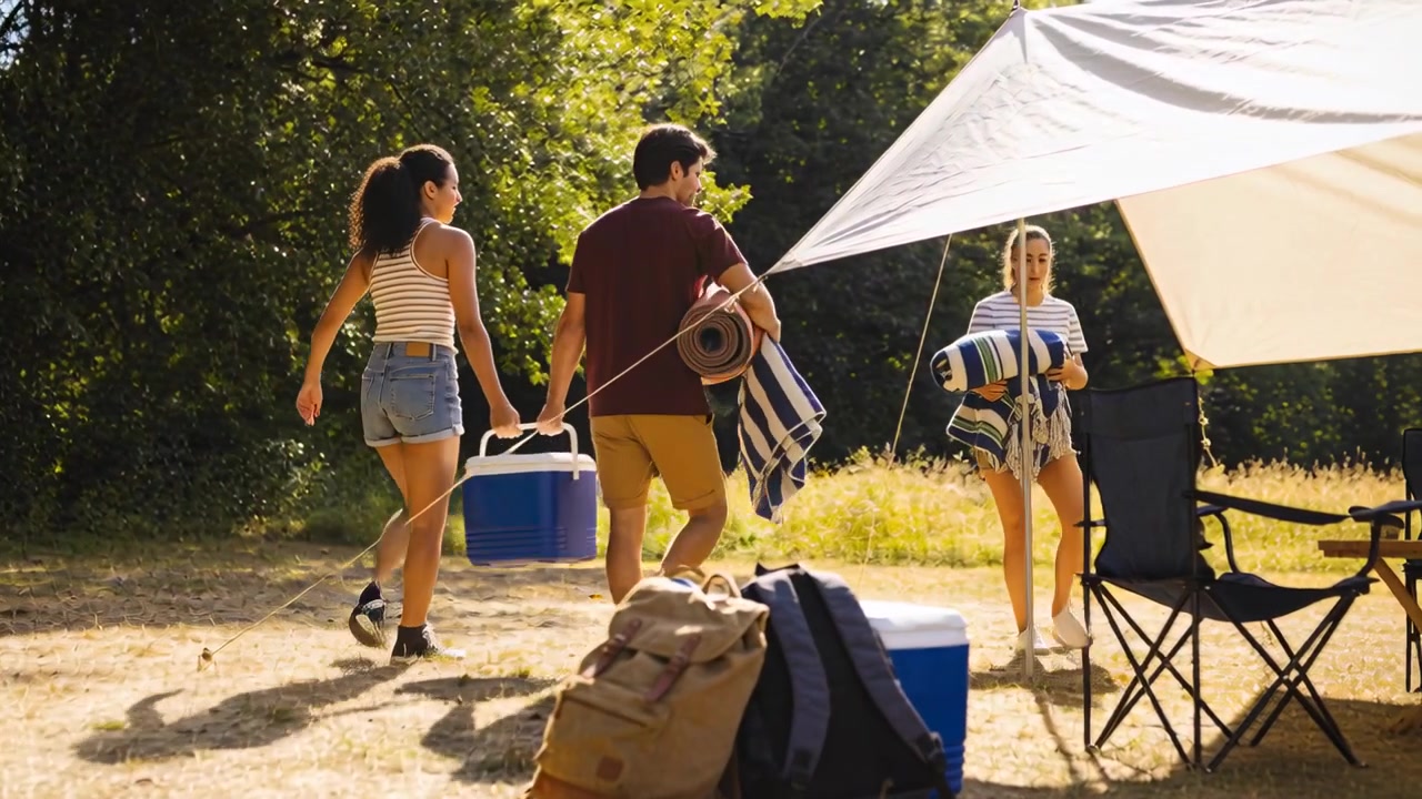 Tre venner, iført stripete overdeler og en burgunderfarget t-skjorte, nærmer seg en baldakin og laster ut en blå kjøleboks og matter.