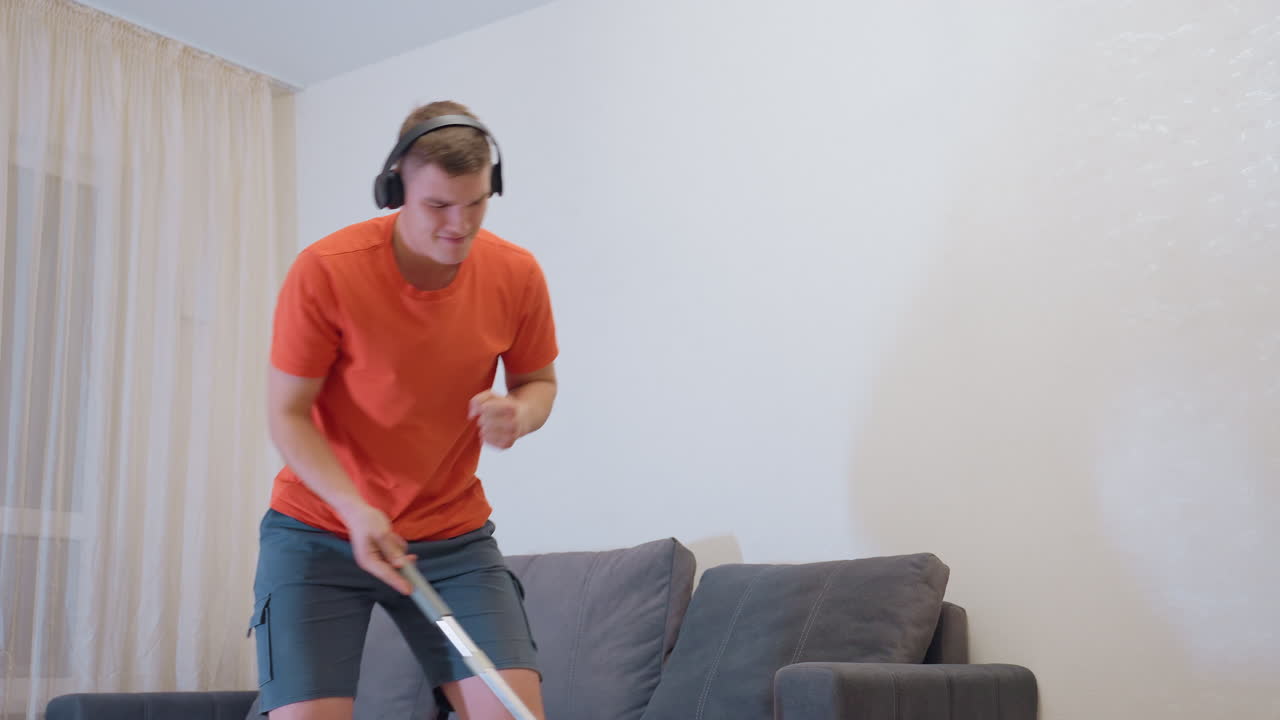 Teen in orange shirt wearing headphones happily dancing while mopping bright living room near sofa enjoying music during cleaning chore with energetic movements making household task fun