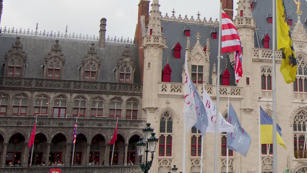 Multiple flags flutter in wind before ornate medieval architecture, overcast daylight, slight camera pan