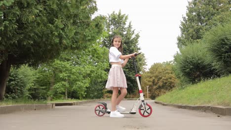 A Little Girl on an Electric Scooter the Wheels of Which Do Not Spin on the Asphalt . A little girl on an electric scooter, the wheels of which do not spin on the asphalt. Trees and plants along the road under a cloudy sky