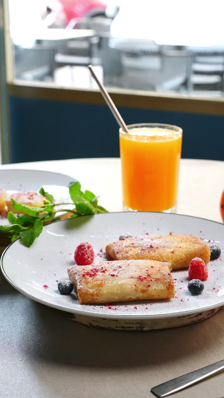 Traditional Ukrainian nalysnyky with forest fruits and powdered sugar are served on a white plate with a glass of fresh orange juice for a healthy breakfast