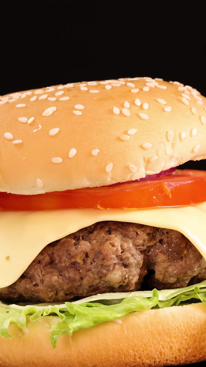 Cheeseburger di manzo succoso con lattuga, pomodoro, cipolla e formaggio fuso che ruota dolcemente su uno sfondo nero sotto un'illuminazione da studio brillante e uniforme.