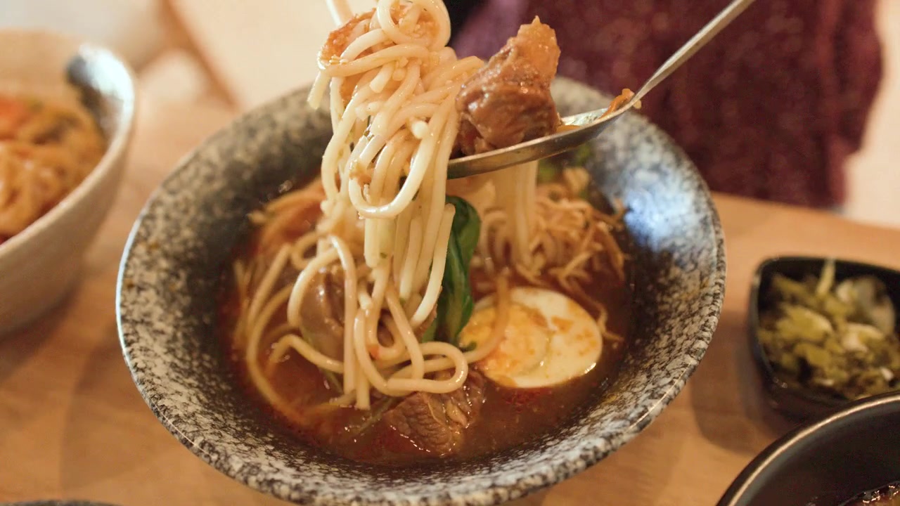 Primo piano di una persona che solleva noodle di manzo birmani con bacchette e cucchiaio a Bangkok, Thailandia.