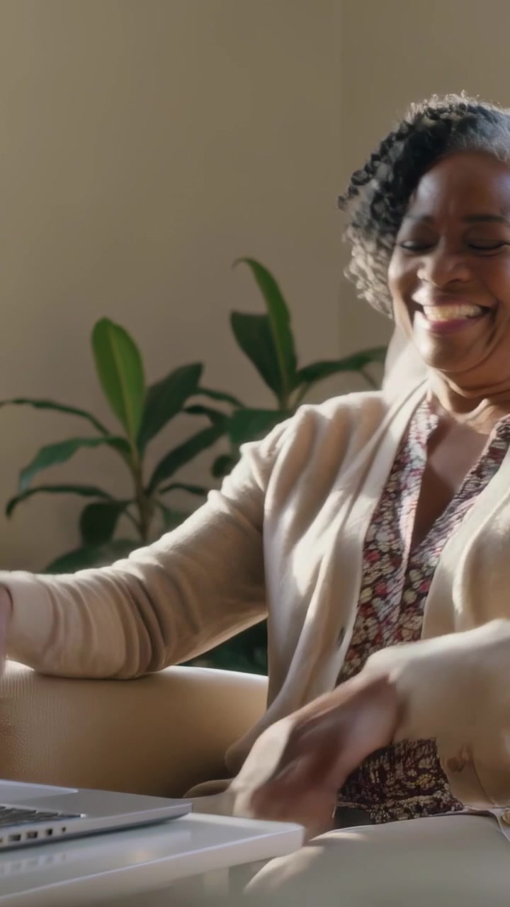 Vertical video: Placing laptop on table, woman in cardigan laughing, reclining in armchair at home