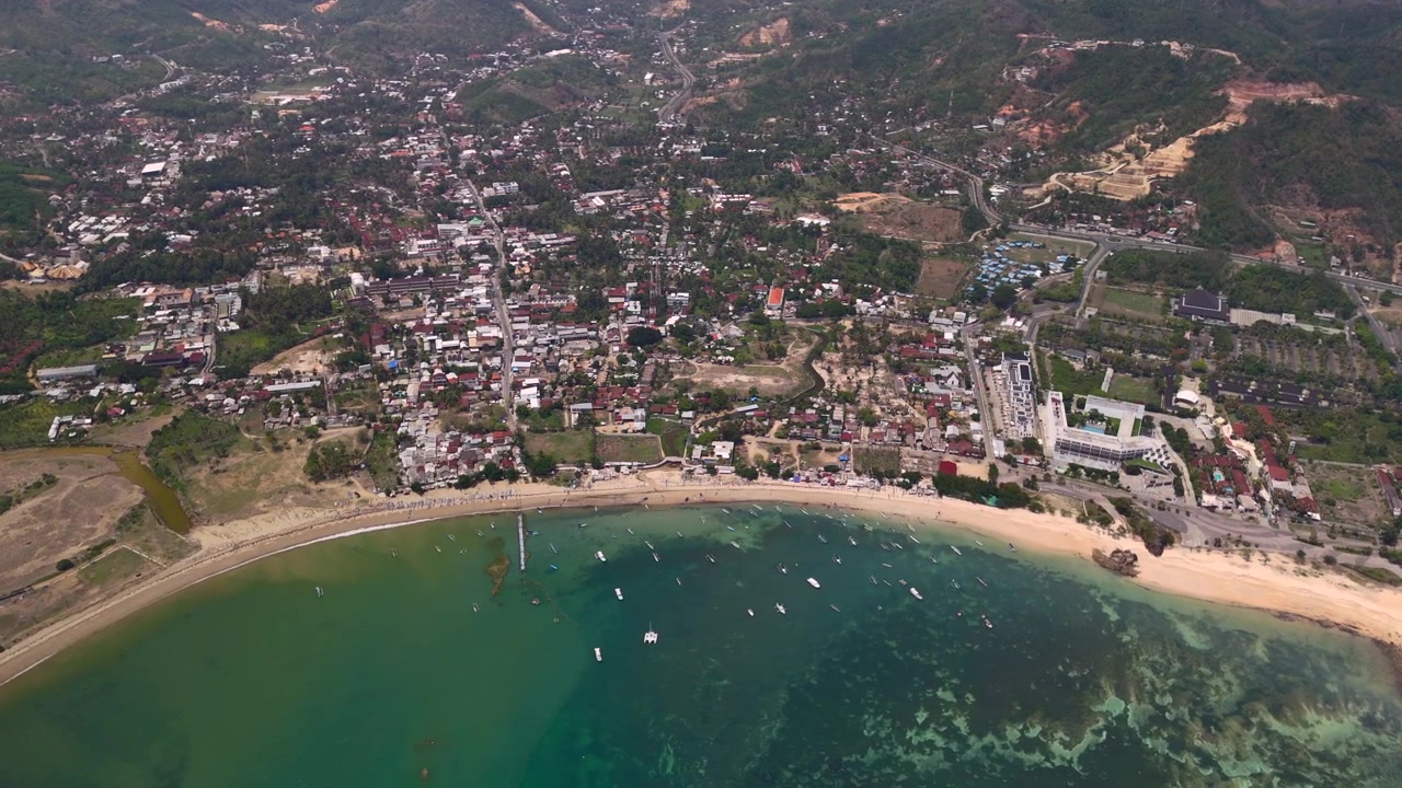 Ripresa con drone della costa di una città in una baia tropicale con barche nell'acqua costiera, colline verdi e case, per temi di viaggio sull'isola, turismo portuale e comunità marittima locale.