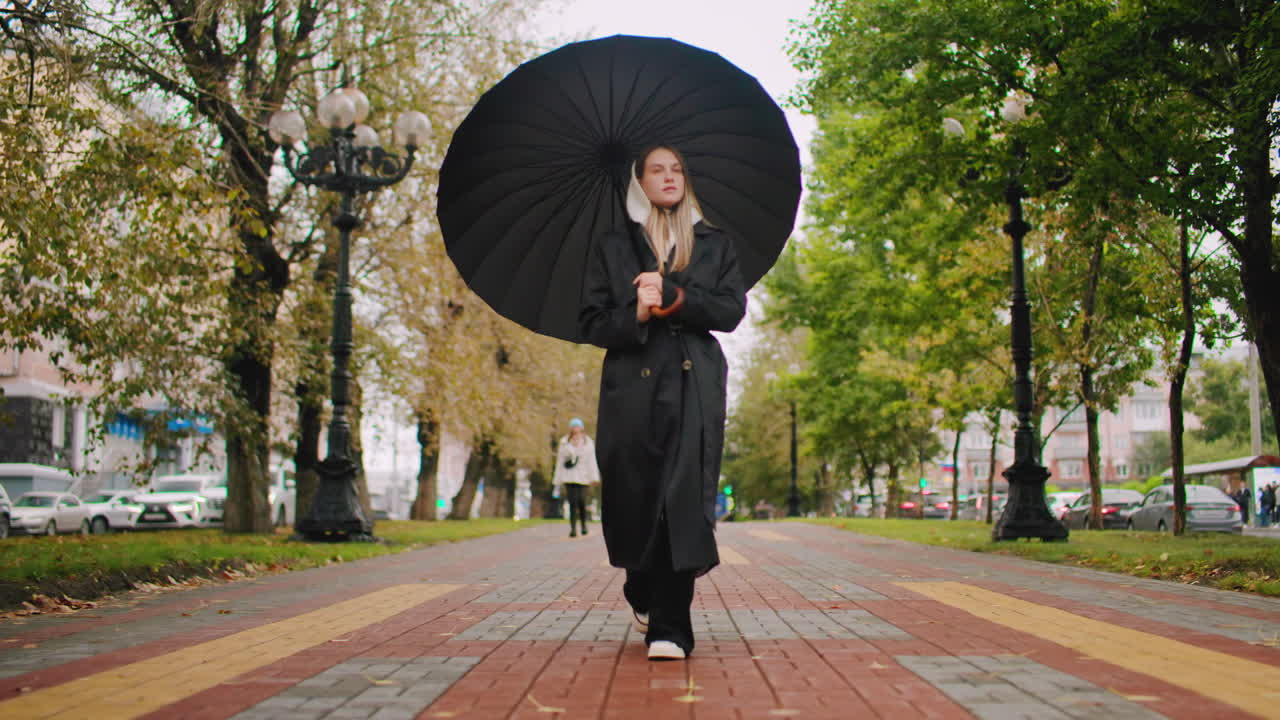 Young woman in long black coat and hood walking alone under umbrella on tree-lined pathway with fallen leaves, cars in background, calm rainy day urban lifestyle scene with moody overcast autumn atmosphere