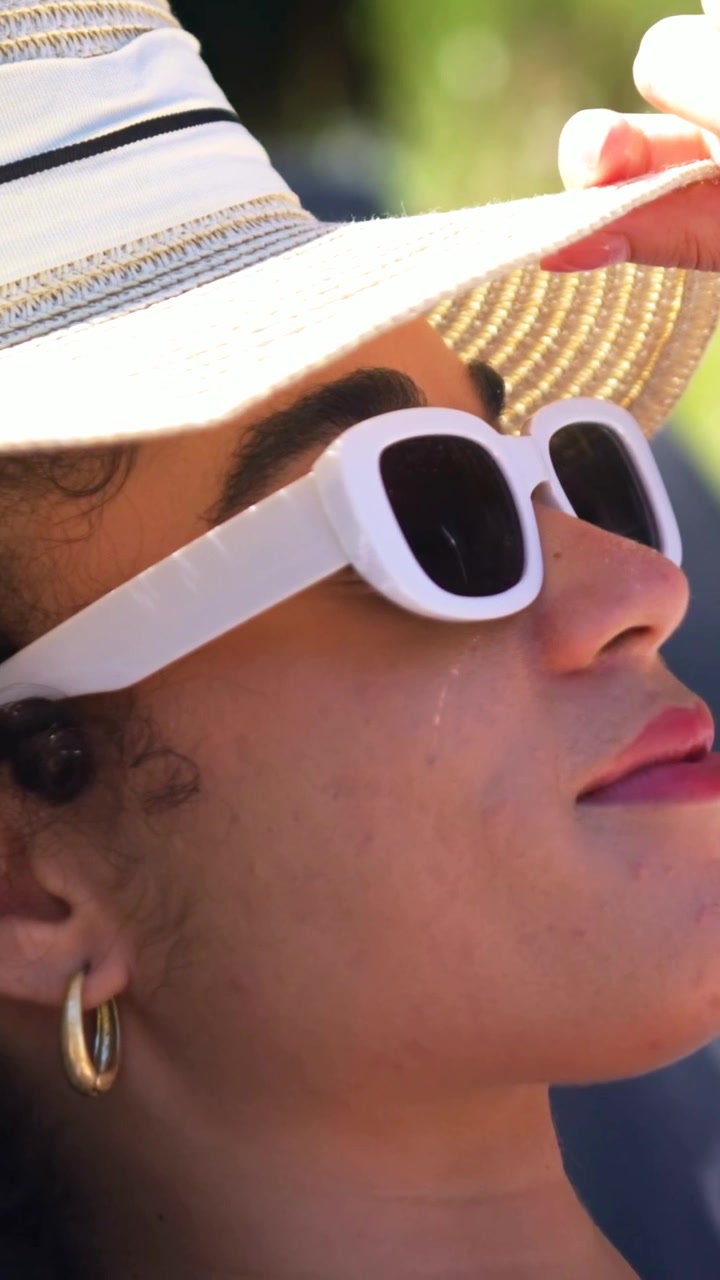 Mujer reclinada protegiéndose los ojos del sol con el ala de un sombrero, la cámara se desplaza hacia arriba mostrando gafas de sol blancas y auriculares de moda.