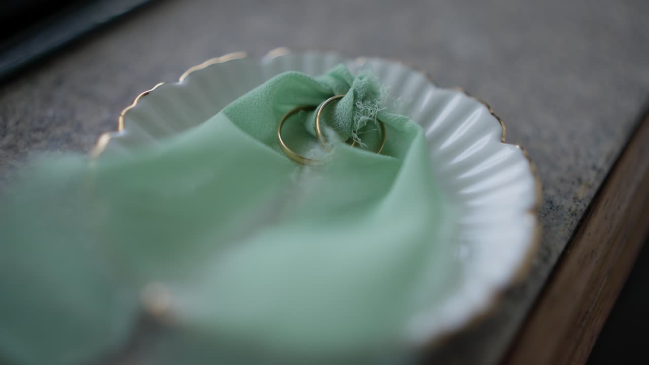 Close up of two gold wedding rings resting on soft green fabric inside a white porcelain dish