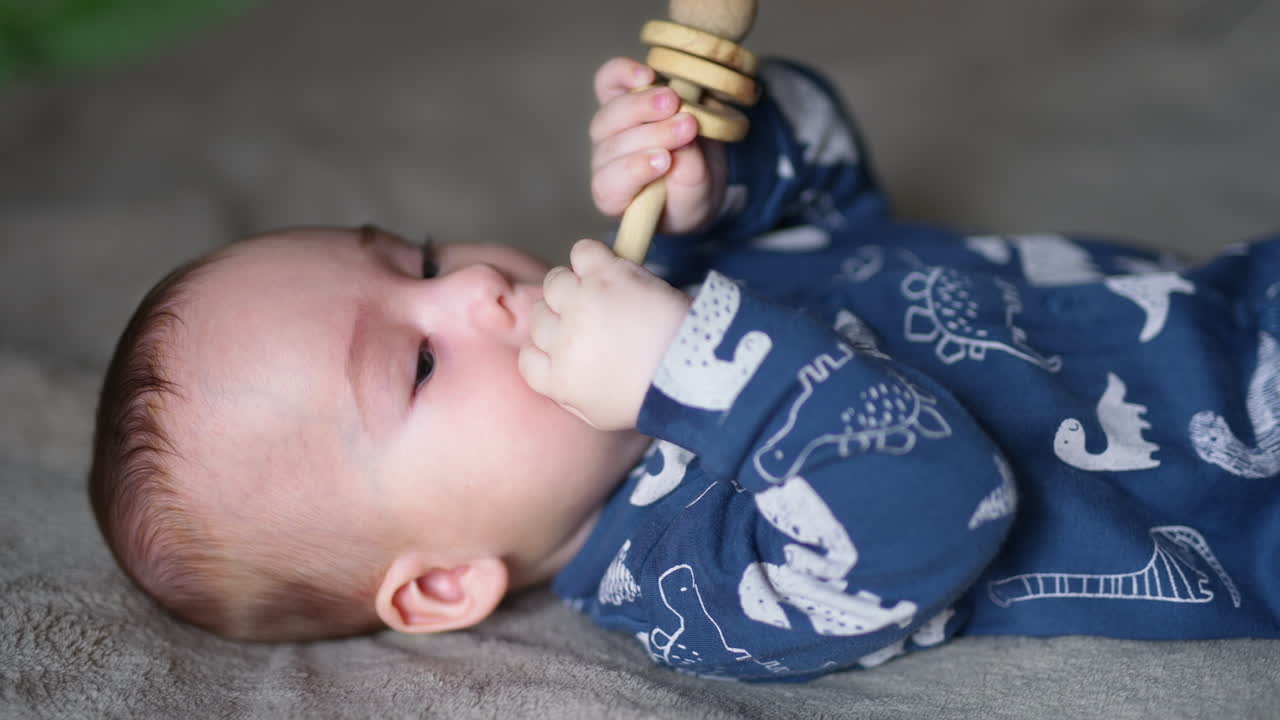 Little boy in blue clothes lies on back holding a wooden toy in hands. Tiny boy puts a toy in his mouth. Close up.