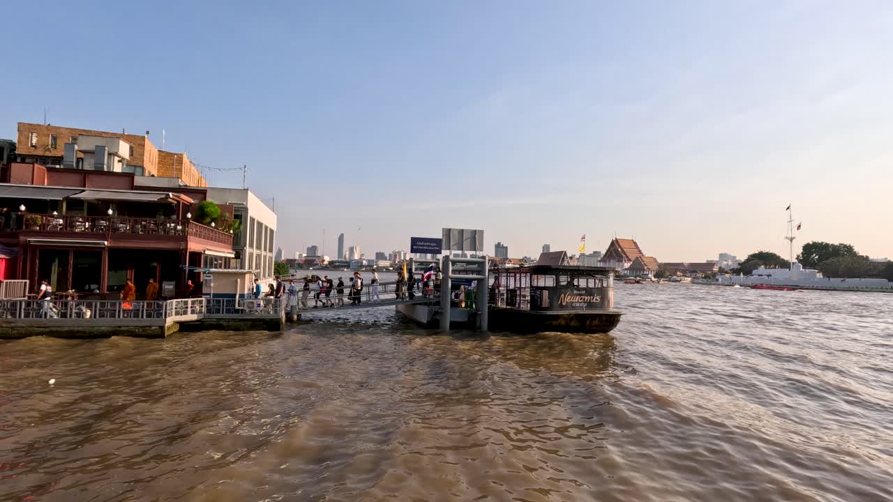 Wide shot of busy Bangkok riverside pier and waterfront restaurant during golden hour river transport