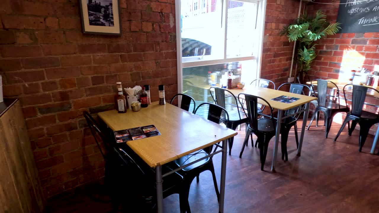A cozy restaurant interior with brick walls, wooden tables, and metal chairs, illuminated by natural daylight through large windows. Subtle camera pan reveals warm, inviting atmosphere