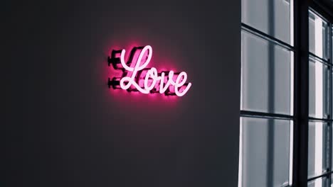 neon pink love sign glowing softly against dark architectural wall, casting diffused light through large window and creating contemporary urban interior mood