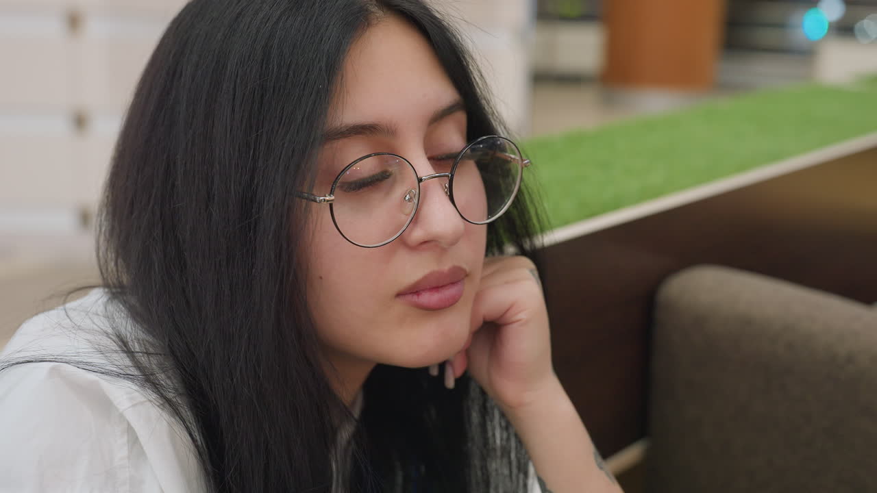 Close up of thoughtful lady adjusting glasses with one hand on jaw, appearing deep in thought while seated indoors with green decor and bokeh blue light softly glowing in background