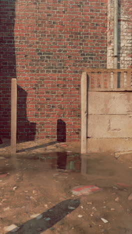 una pared de ladrillo con una valla de hormigón delante de ella. hay un charco en el suelo y algunas tuberías de metal oxidadas.