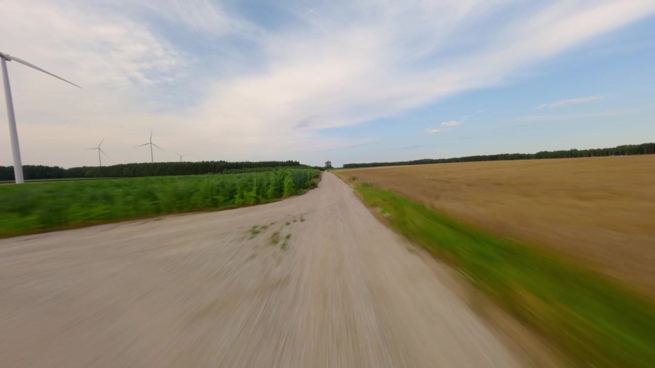 Volo veloce a bassa quota sopra una strada di campagna tra campi di grano e mais con turbine eoliche sullo sfondo. Concetto di sostenibilità ed energia ecologica.