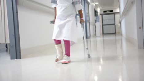 Lowsection-of-african-american-girl-in-hospital-gown-walking-using-crutches-in-corridor,-slow-motion