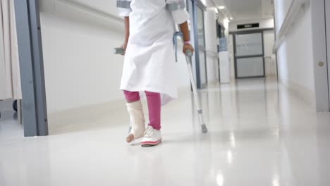 lowsection of african american girl in hospital gown walking using crutches in corridor, slow motion