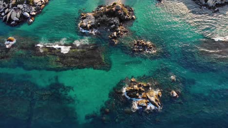 Aerial-view-of-rocky-coast,-a-beach-and-shallow,-turquoise-sea,-sunny-day,-in-Tayrona-National-Park,-in-the-Caribbean-region-of-Colombia---Tilt-up,-drone-shot