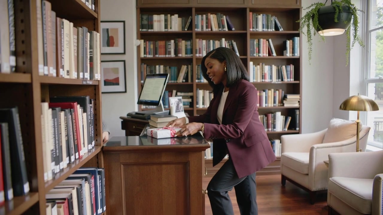 Leaning across library desk, woman in maroon blazer handing wrapped package to staff for check-in