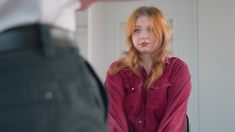 close up of young woman in red shirt seated indoors with neutral expression, looking forward attentively at person in front of her who is partially visible