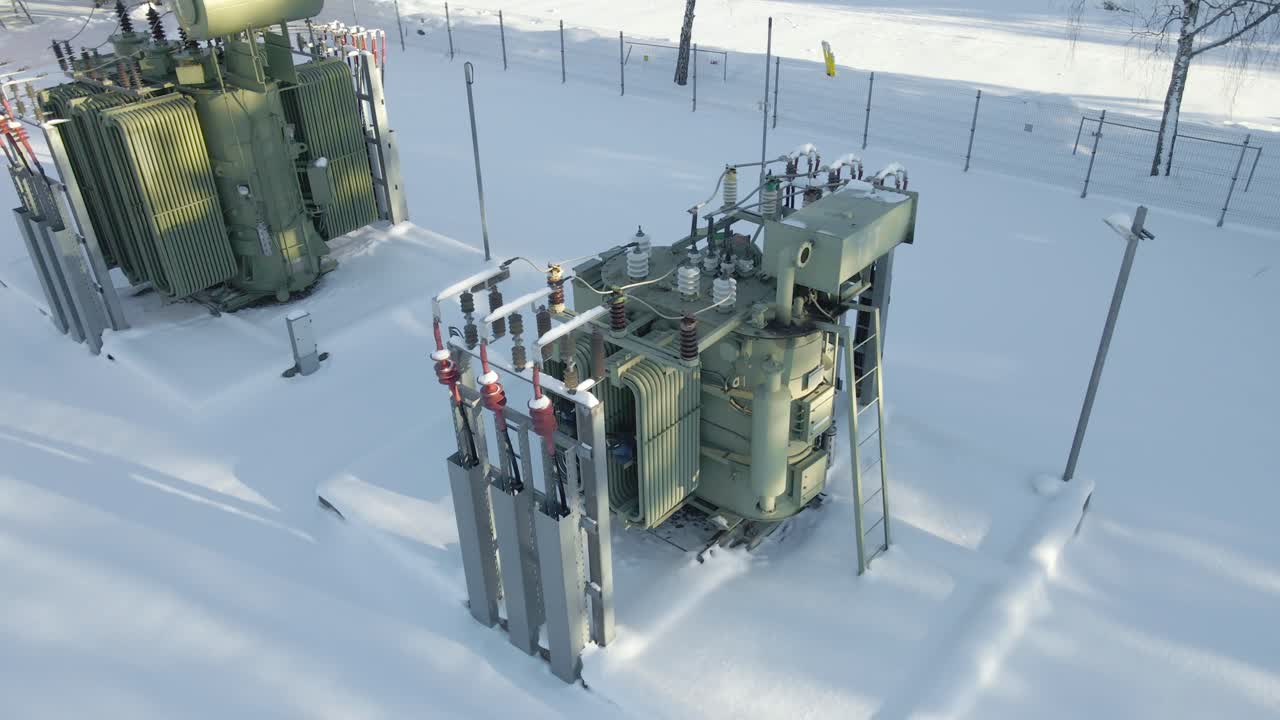 Cinematic aerial descent reveals snow covered power transformers with cooling fins and high voltage bushings, standing in a frozen electrical substation under bright winter sunlight