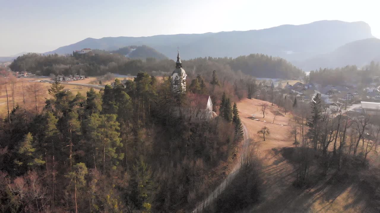 Luftaufnahme einer Kirche, eingebettet in den Bergen Sloweniens. Die Kamera bewegt sich vom Boden zum Himmel und blickt über die Kirche in den Alpen.