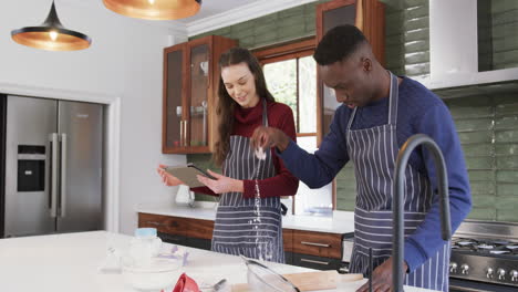happy diverse couple standing in kitchen, using tablet and sprinkling flour on worktop,slow motion