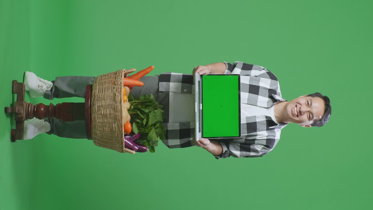 полное тело азиатского мужчины-фермера с корзиной с овощами, показывающей зеленый экран ноутбука на зеленом экране на заднем плане в студии