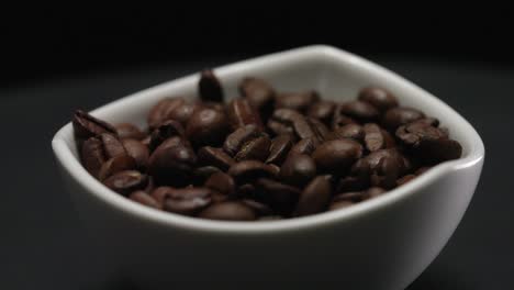 sow-motion close up of roasted coffee beans falling in a white ceramic dish rotating gracefully on a platform. captures the texture and richness of coffee.