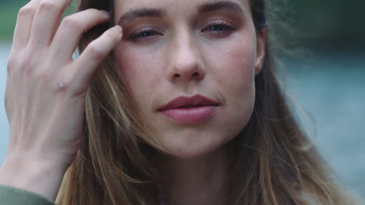 retrato de una joven independiente que parece confiada y con expresión seria pasando la mano por el cabello con el viento soplando al aire libre junto al lago 4k