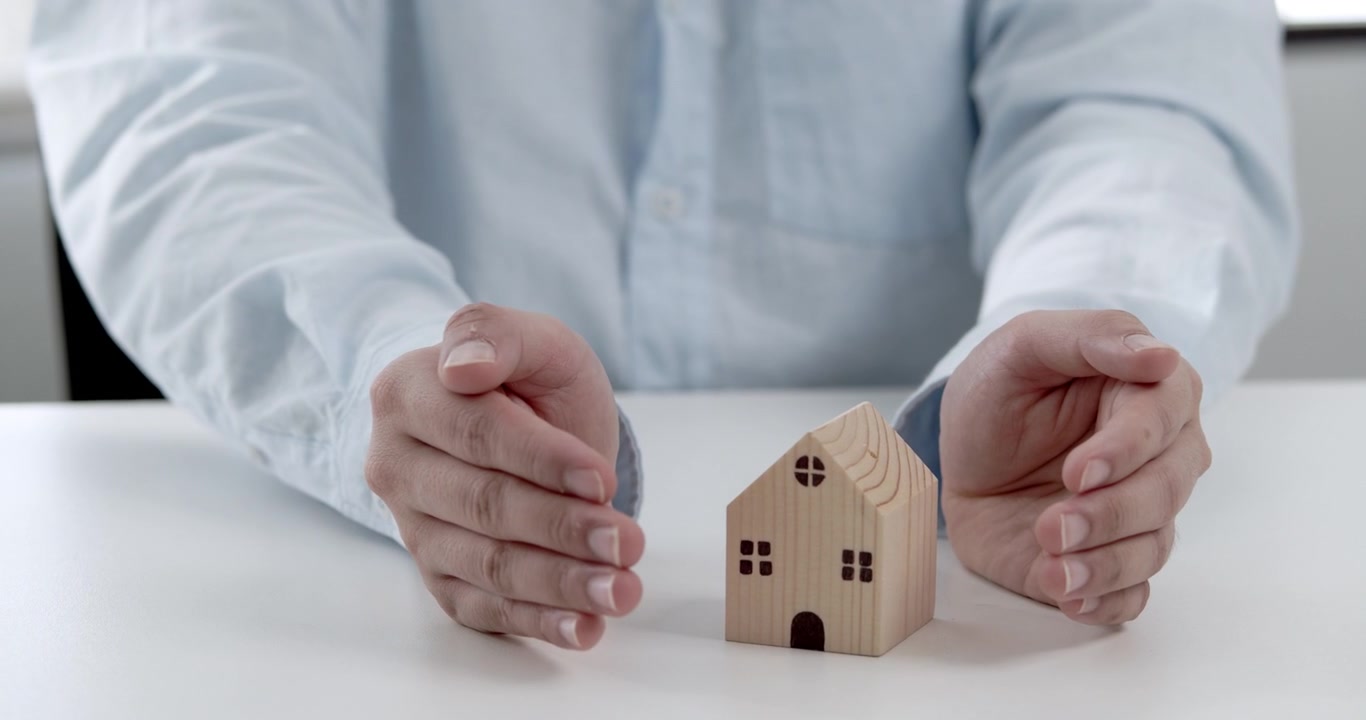Las manos de un joven con camisa azul claro sosteniendo una maqueta de casa de madera, evocando protección cálida para el hogar y seguros.