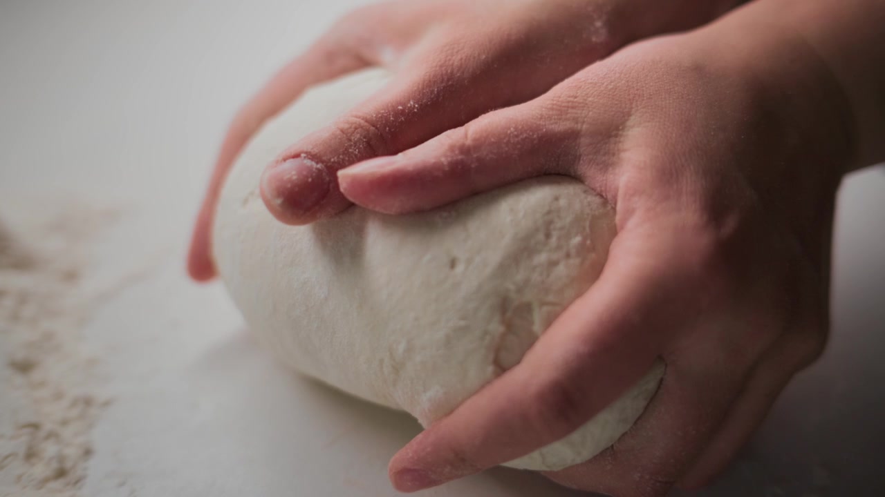 Primo piano di mani che impastano l'impasto su una superficie infarinata. Le dita premono e piegano l'impasto con cura. La messa a fuoco morbida enfatizza la consistenza e il movimento. Ideale per contenuti di cucina, panificazione o preparazione di alimenti