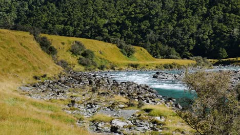 Wunderschöner-Fluss,-Der-Bei-Sonnenlicht-Zwischen-Bewachsenen-Bergen-Und-Grünen-Feldern-Fließt---Schwenkaufnahme