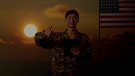 asian man soldier smiling and pointing to a tablet in his hand while standing with flag of the united states, sunset time