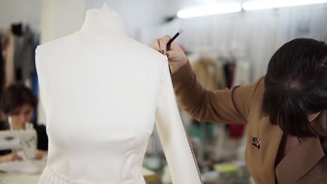 modern sewing studio, concentrated brunette female fashion designer measuring sleeve from a white suit on mannequin and making notes holding her notebook. slow motion