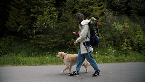 A-man-of-tourists-in-special-clothes-for-a-hike-with-a-large-backpack-with-his-light-colored-dog-walks-along-the-forest-road