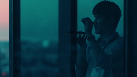asian businessman using smartphone talking to client corporate executive negotiating deal having phone call working late in office looking out window at sunset