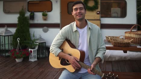 El-Hombre-Toca-La-Guitarra,-Sostiene-La-Guitarra-Acústica-Y-Sonríe-A-La-Cámara.-Casa-Sentada-Contra-Las-Ruedas,-Remolque-Con-Bonita-Decoración-Exterior-Alrededor.-Retrato