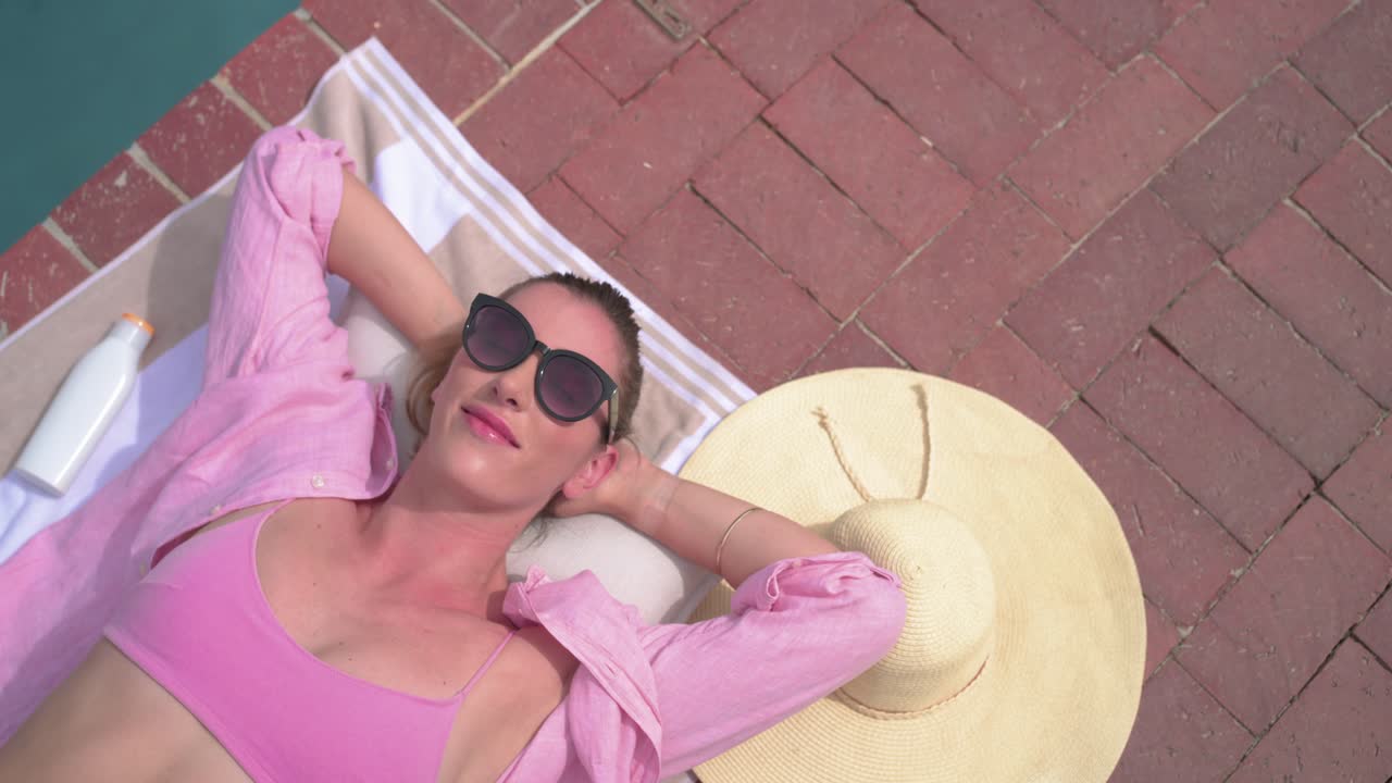 Camera panning revealing adult female lying on towel poolside, smiling, sunbathing in pink swim top