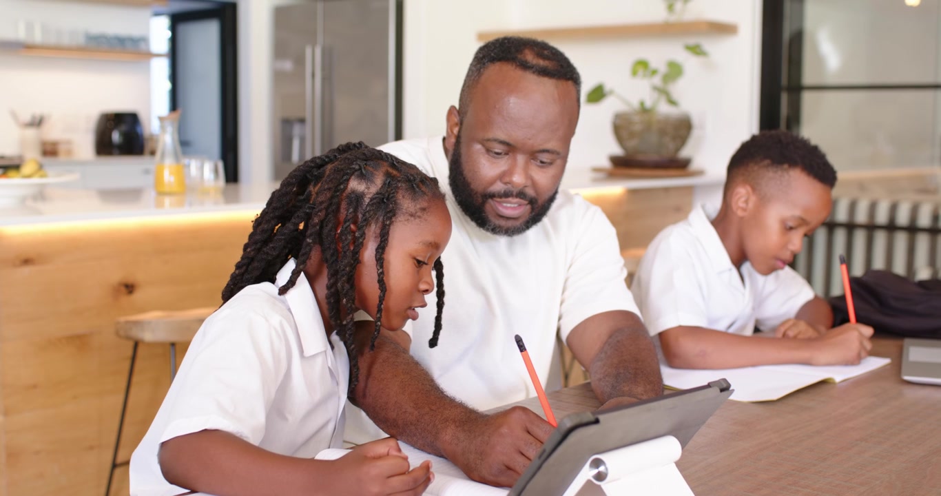 African American dad teaching two boys at kitchen table, pointing tablet while left child speaking