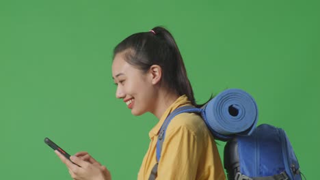 close up side view of asian female hiker with mountaineering backpack smiling and looking at smartphone while walking on green screen background in the studio