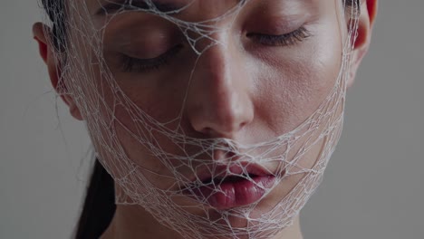 white mesh veiling delicate features, young woman posing with closed eyes in studio portrait, embodying vulnerability, identity concealment, and emotional depth against minimalist gray background