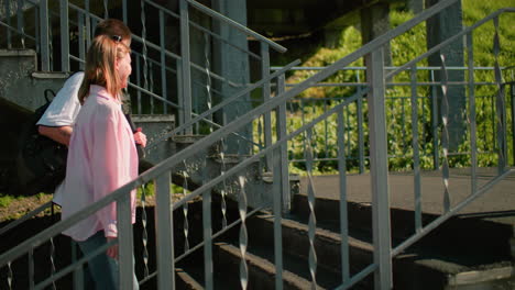 students climbing outdoor staircase in urban setting, interacting warmly while carrying backpacks, sunlight casting shadows on metal railings, capturing casual conversation and connection