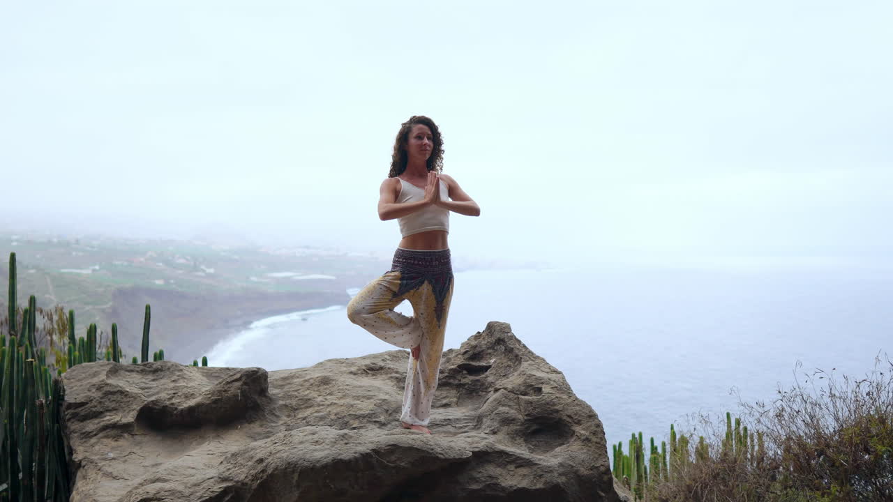 en una isla en medio de las montañas, una mujer joven practica yoga, de pie sobre una pierna, brazos levantados, con vistas al océano