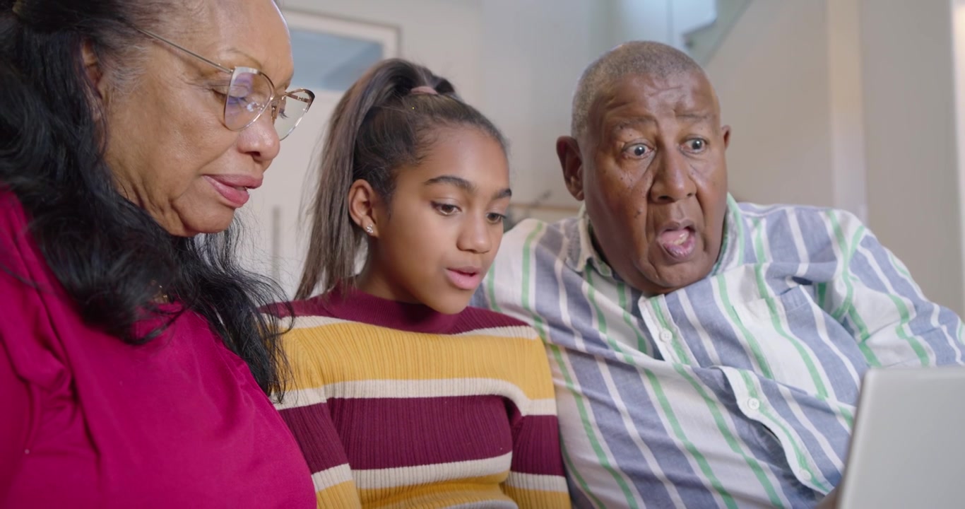 African American grandparents watching teen granddaughter in striped top explaining laptop on couch